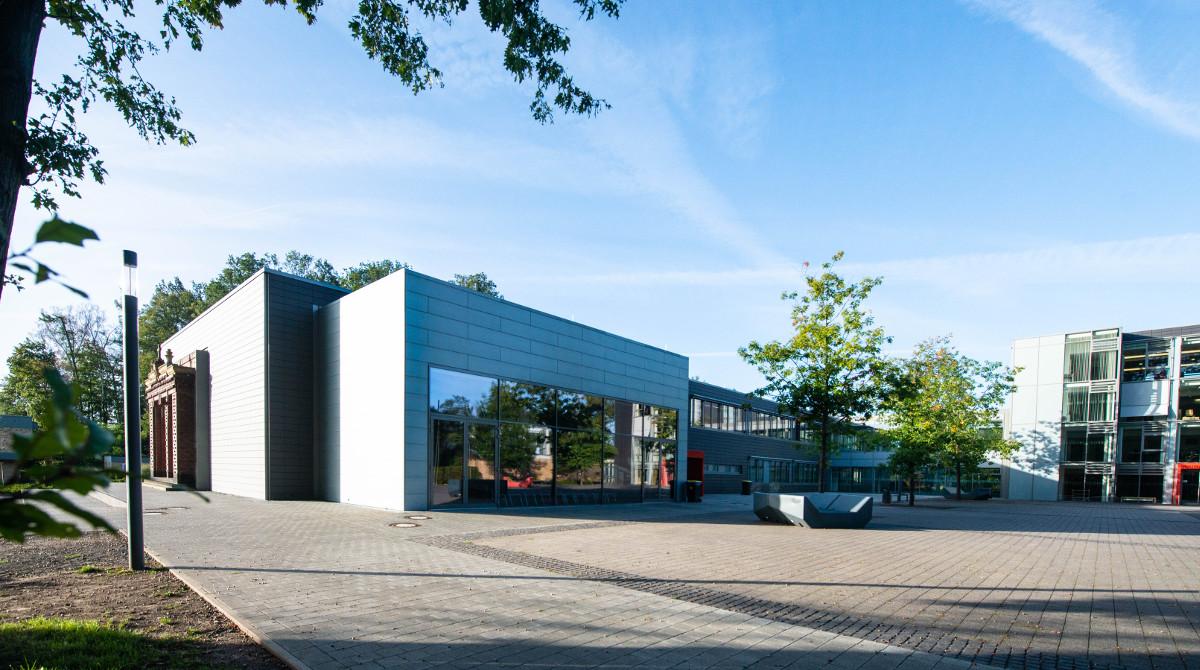 Modern gray building with large windows and tree-lined paved courtyard under blue sky