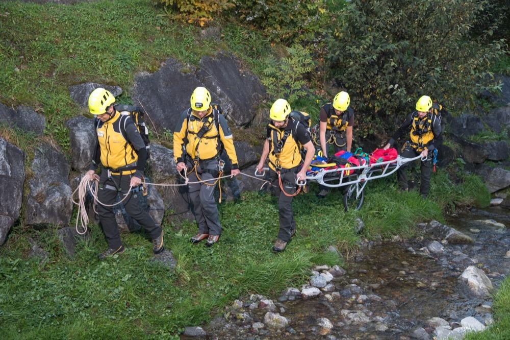 Five mountain rescuers in yellow helmets carrying a stretcher with a patient along a rocky stream