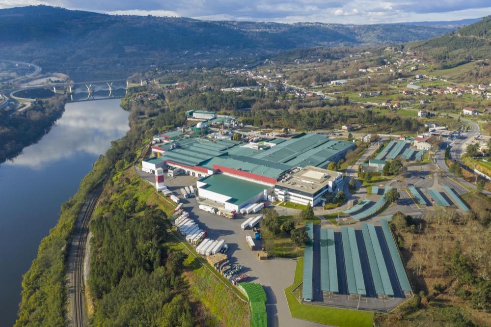 Aerial view of a large industrial facility with green roofs beside a river and rolling hills.