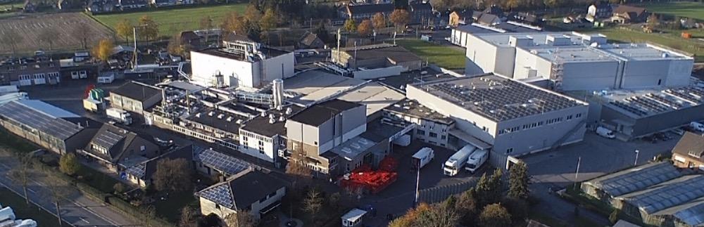 Aerial view of an industrial complex with white warehouses, solar panel roofs, delivery trucks and surrounding fields.