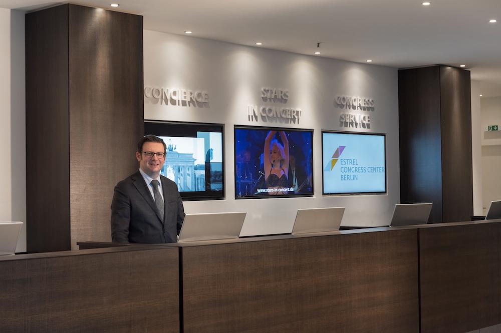 Modern wooden reception desk with a smiling concierge in suit and three digital information screens behind.