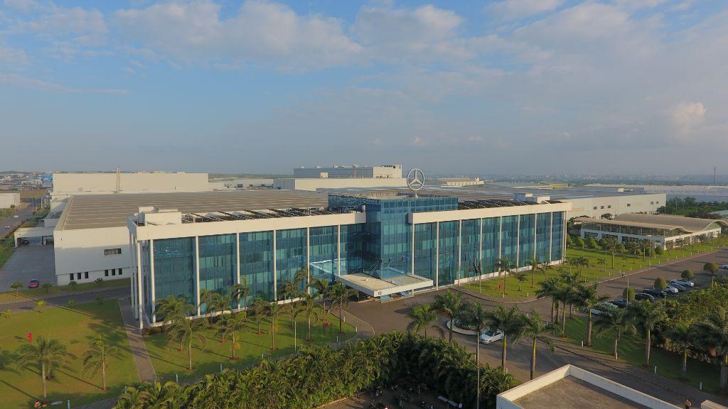 Aerial view of a modern Mercedes-Benz factory building with glass facade, rooftop logo and palm-tree lined grounds.
