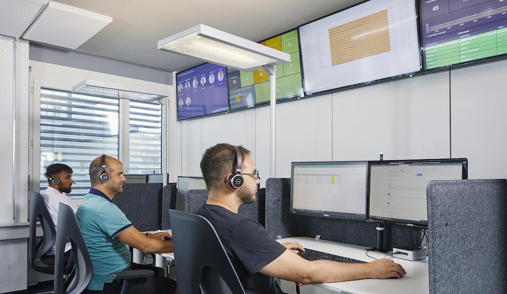 Three office employees seated at desks wear headsets and work on computers with multiple monitors.