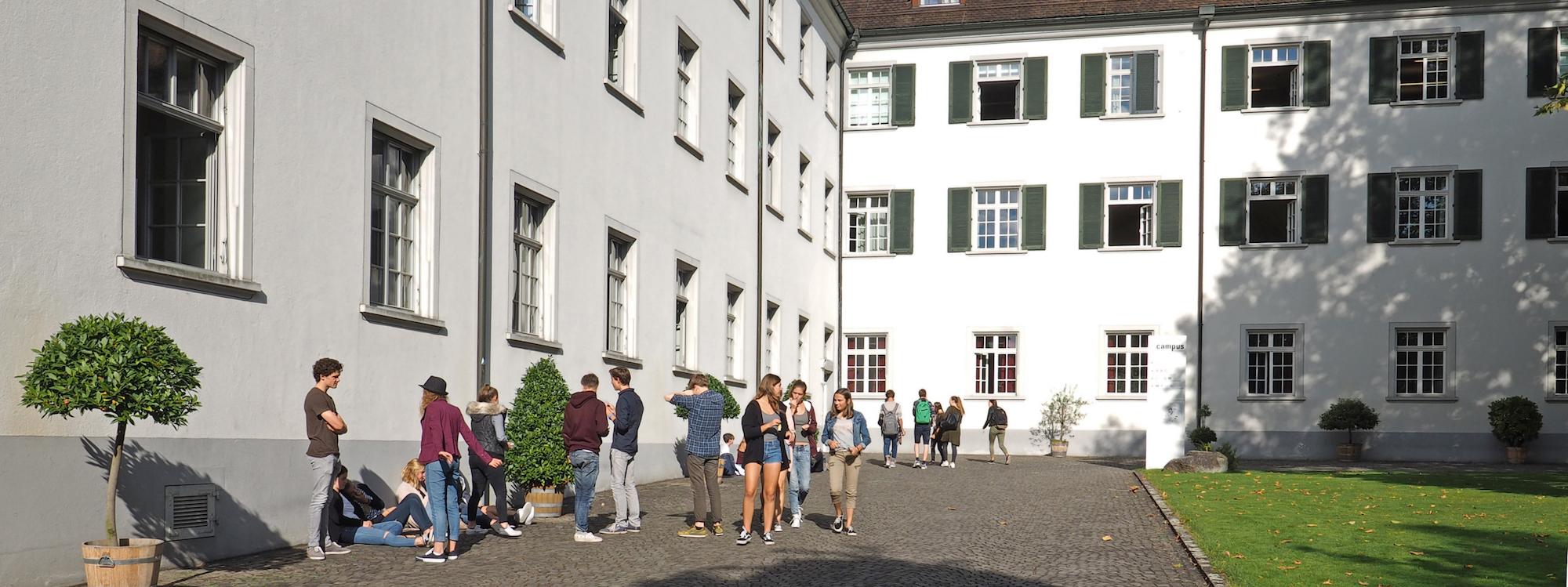 Students chatting and walking in sunny courtyard beside white campus building