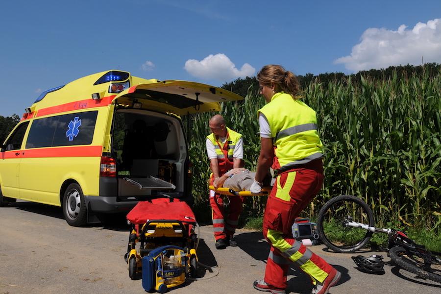 Two paramedics loading an injured cyclist on a stretcher into a yellow ambulance by a country road