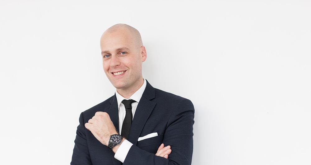 Male model in black suit displaying a black chronograph wristwatch on his left wrist against white background