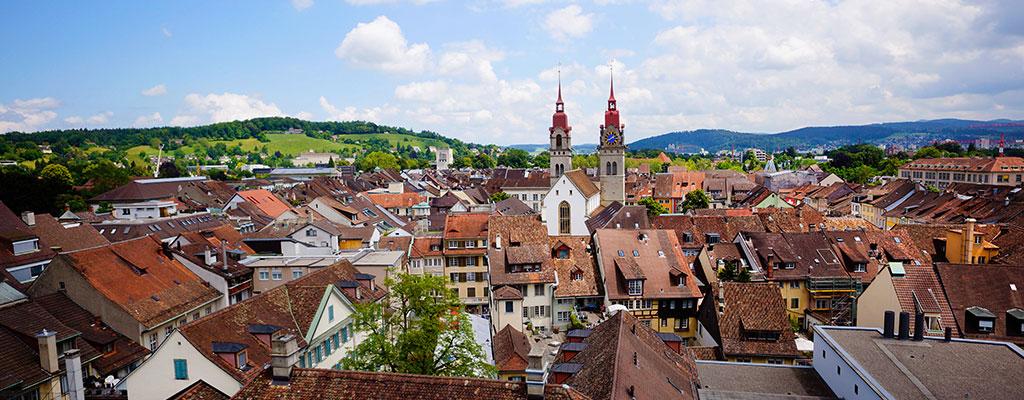 Panoramic view of a European town with red-tiled roofs and a twin-spired church beneath a cloudy sky