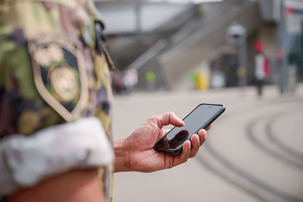 Person in military clothing interacting with a smartphone next to train tracks.