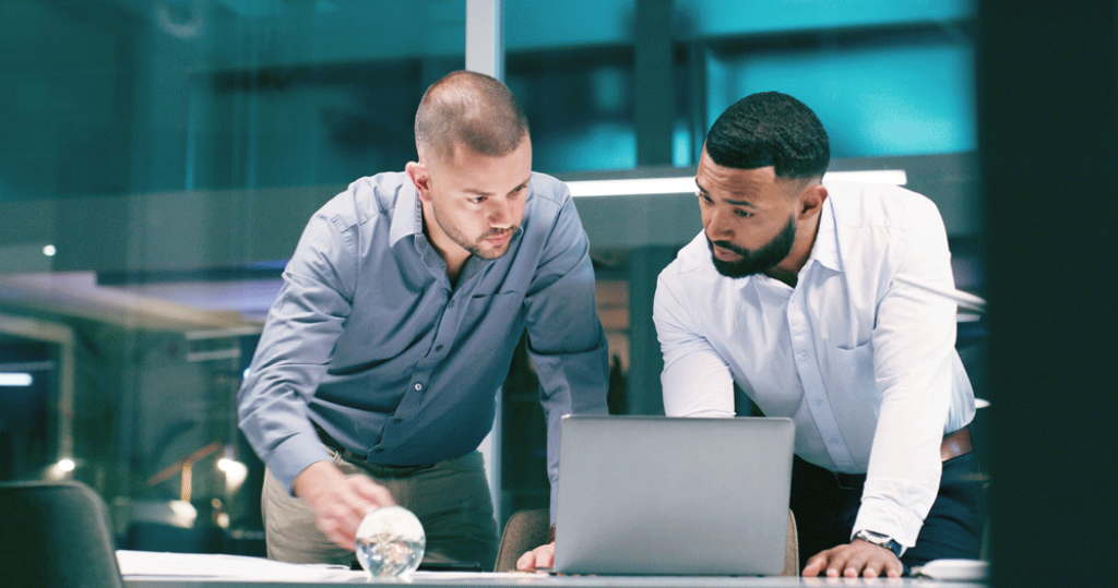 Two men in professional clothin in an office environment focused on a laptop.