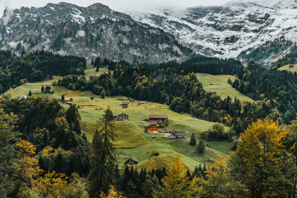 Swiss alpine village with wooden chalets on green meadows below snow-capped peaks in fall.