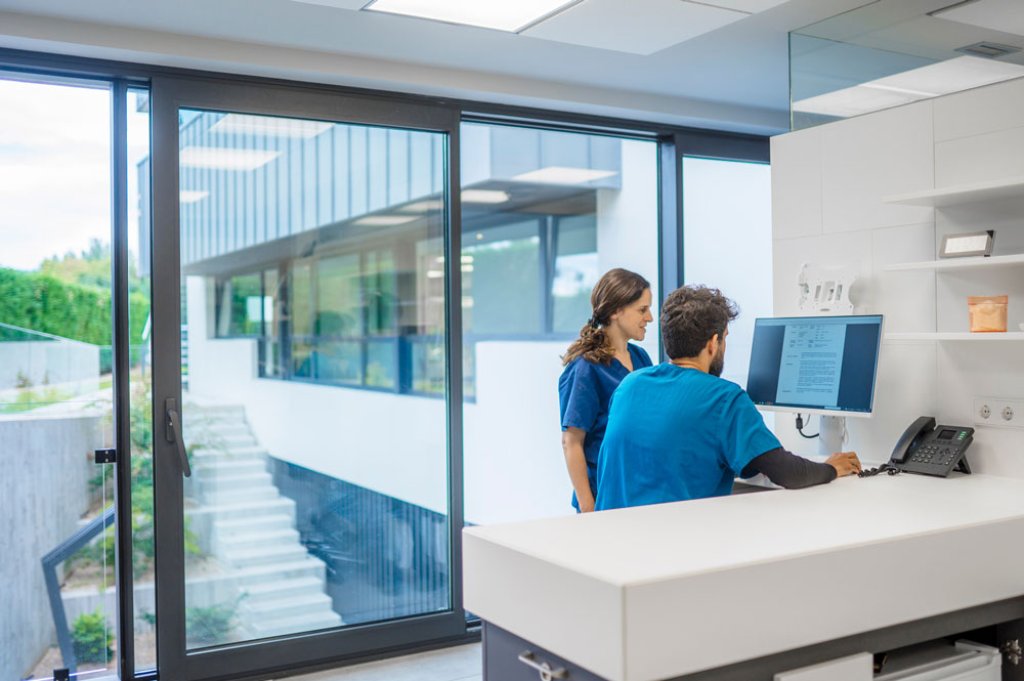 Two healthcare professionals in blue scrubs working at a computer in a modern medical office with large windows.