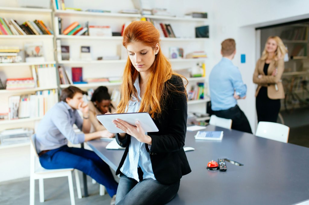 Frau, die in einem modernen Lehrerbüro ein Tablet benutzt, während Kollegen im Hintergrund arbeiten.
