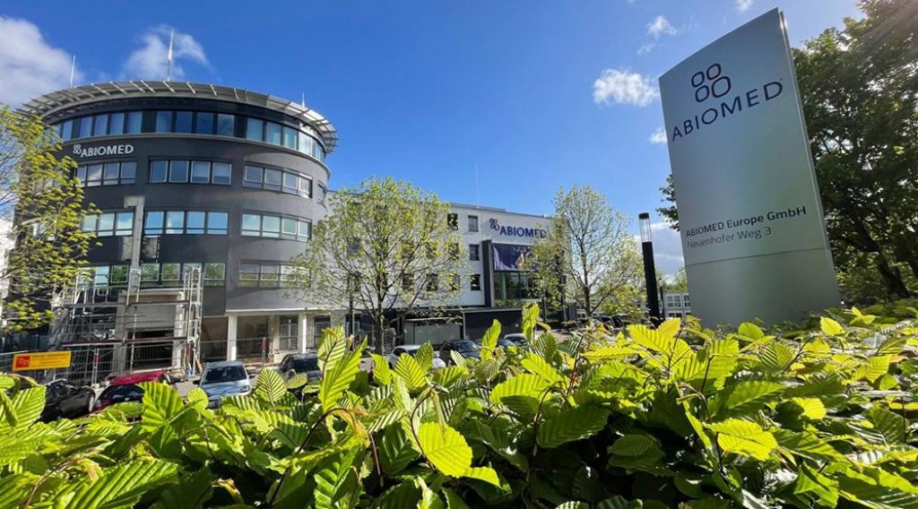 Exterior view of Abiomed Europe GmbH&rsquo;s modern curved office building and signpost behind green hedge under blue sky.