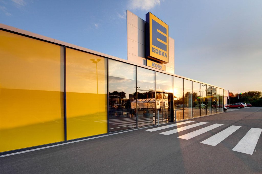 Exterior of modern Edeka Kolb supermarket with yellow panels, glass entrance, and zebra crossing in front.