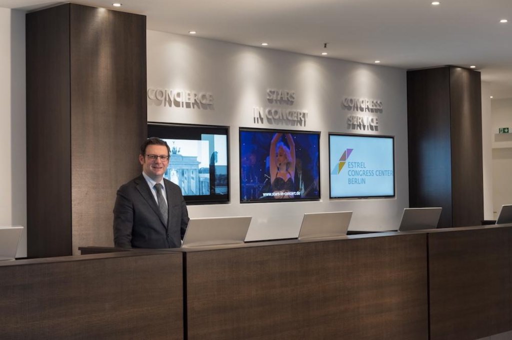 Modern wooden reception desk with a smiling concierge in suit and three digital information screens behind.