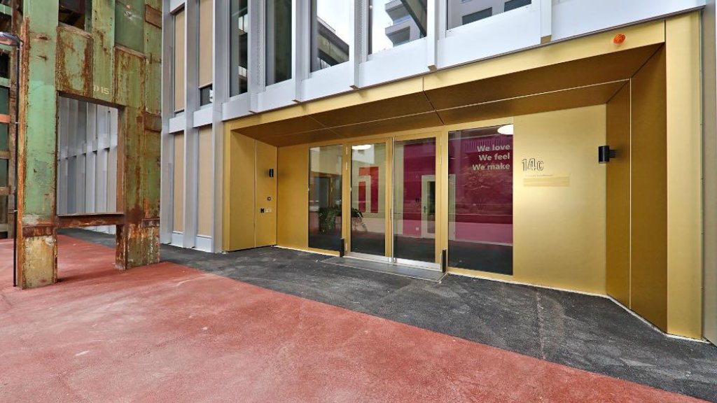 Entrance of a modern building with gold facade, glass doors, rusted metal structure and red pavement.