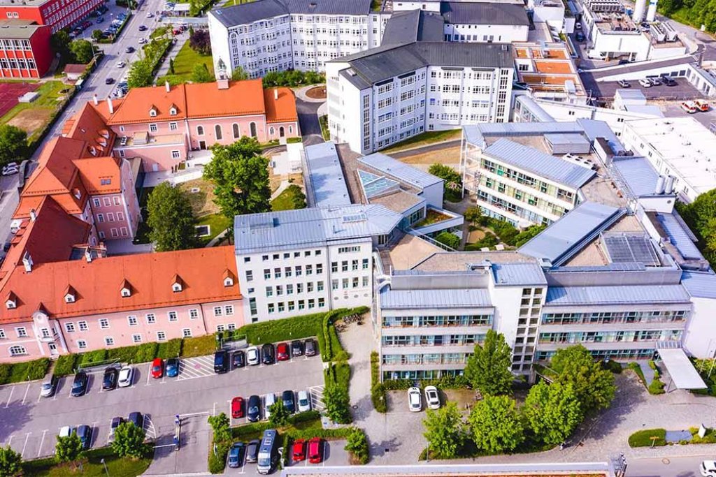 Luftaufnahme von verbundenen modernen und historischen Geb&auml;uden mit roten D&auml;chern, Parkplatz und B&auml;umen.