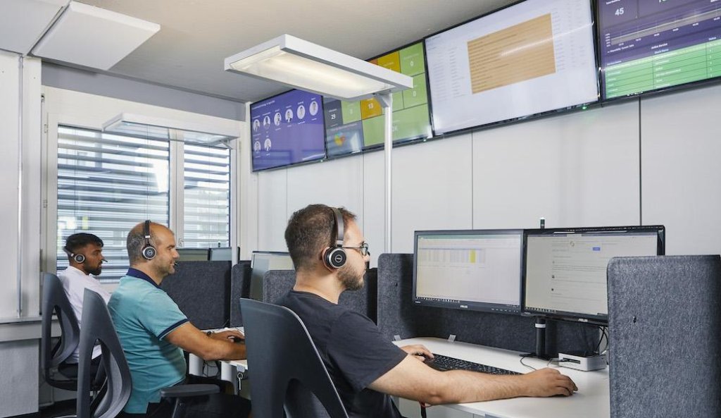 Tres empleados de oficina sentados en escritorios llevan auriculares y trabajan en ordenadores con varias pantallas.