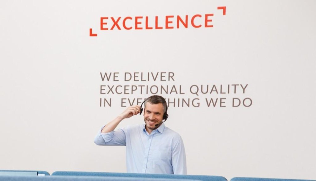 Agente de atenci&oacute;n al cliente sonriente con auriculares frente a pared de oficina con letrero &laquo;EXCELLENCE&raquo;.