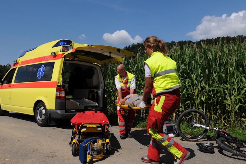 Zwei Sanit&auml;ter heben einen verletzten Radfahrer auf eine Trage und laden ihn in einen gelben Rettungswagen.