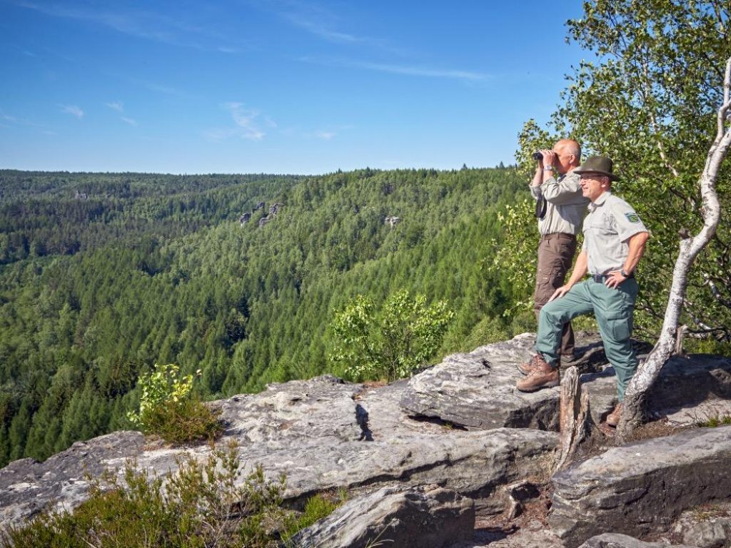 Deux gardes forestiers sur une falaise rocheuse observant une for&ecirc;t dense, l&rsquo;un utilise des jumelles.
